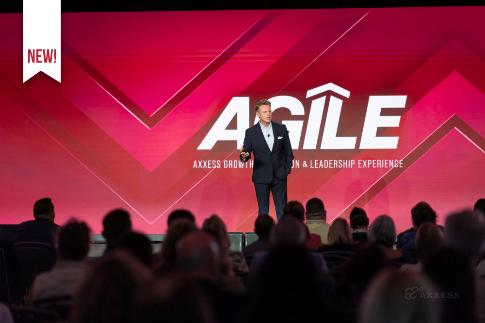 A man in a suit is standing on a stage in front of a large screen with the word "AGILE" on it. He is speaking to a large audience.