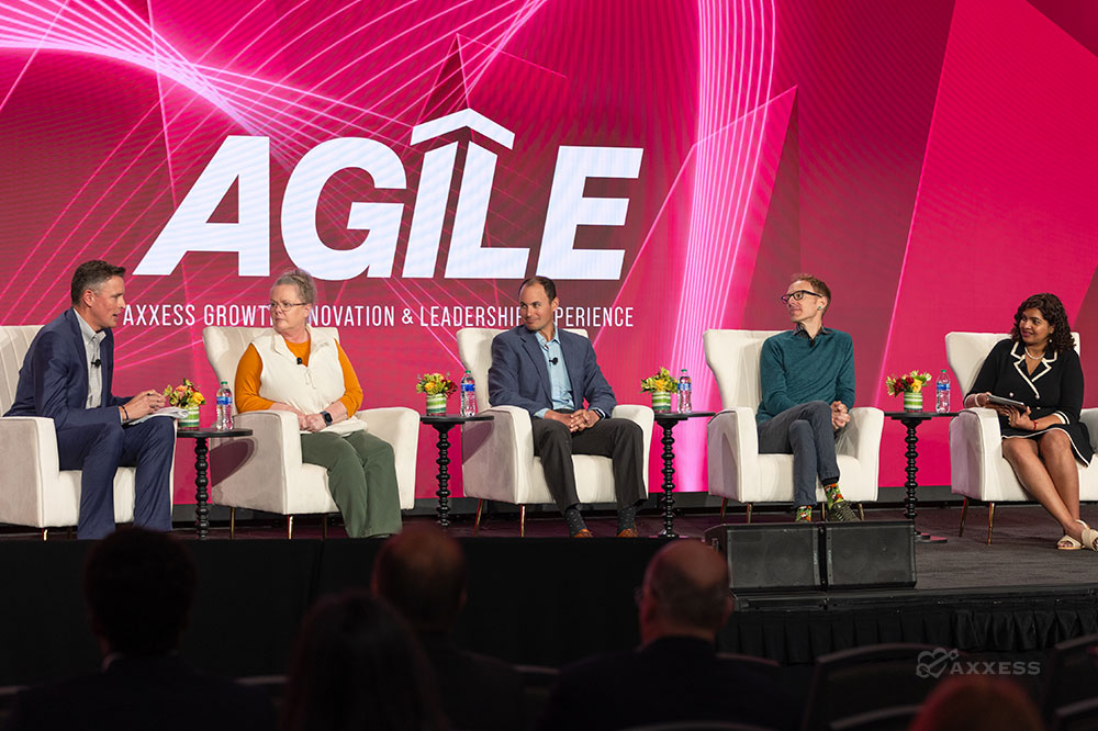 A panel discussion at a conference. There are five people on the panel, four men and one woman. They are all sitting in chairs on a stage. There is a large screen behind them that says "Agile."